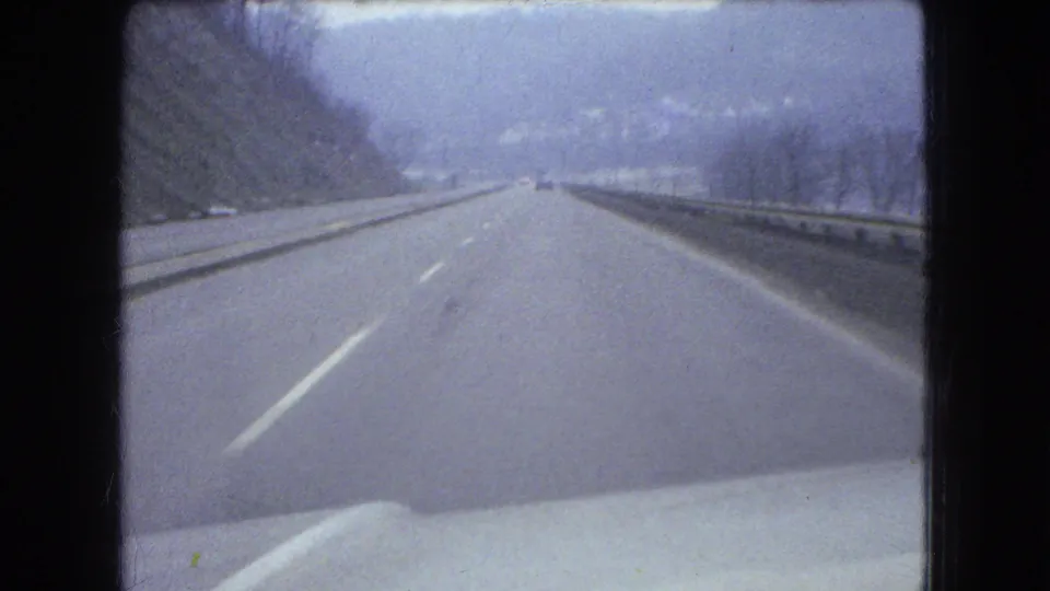 1966 Ohio 8mm forward-driving view on a wet road, with lane markings, guardrails, bare roadside trees, hills in haze, and the vehicle hood visible along the bottom.