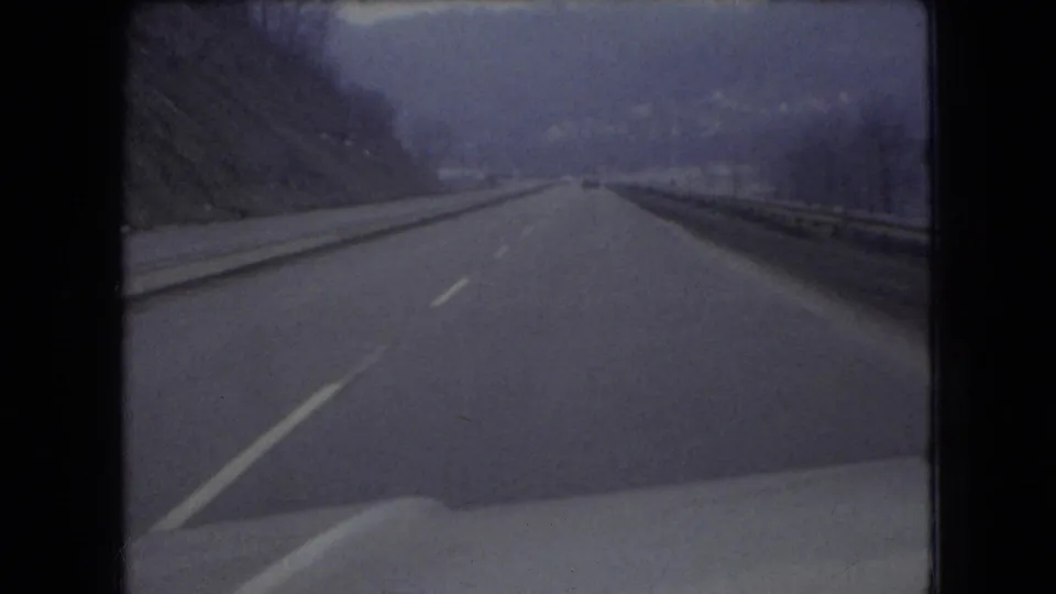 1966 Ohio 8mm frame looking ahead from inside a moving vehicle, showing wet pavement, a center line, roadside guardrail, distant traffic, hills, and the car hood in the foreground.