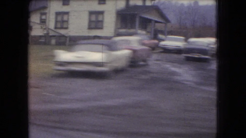 1966 Ohio home-movie frame showing a two-story house, several mid-century cars, and wet or muddy ground viewed in motion.