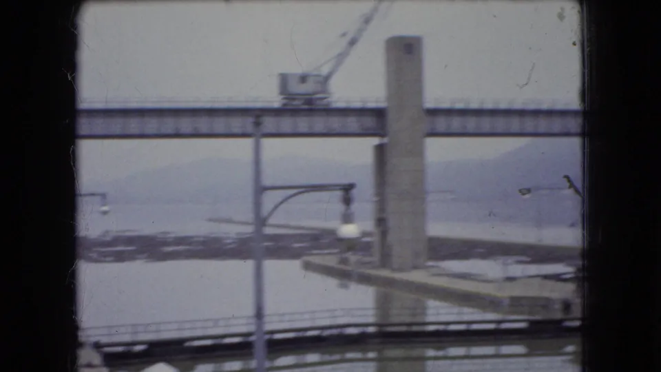 1966 Ohio construction view showing a concrete bridge pier, elevated span, crane, and distant water or landscape through dark film or window edges.