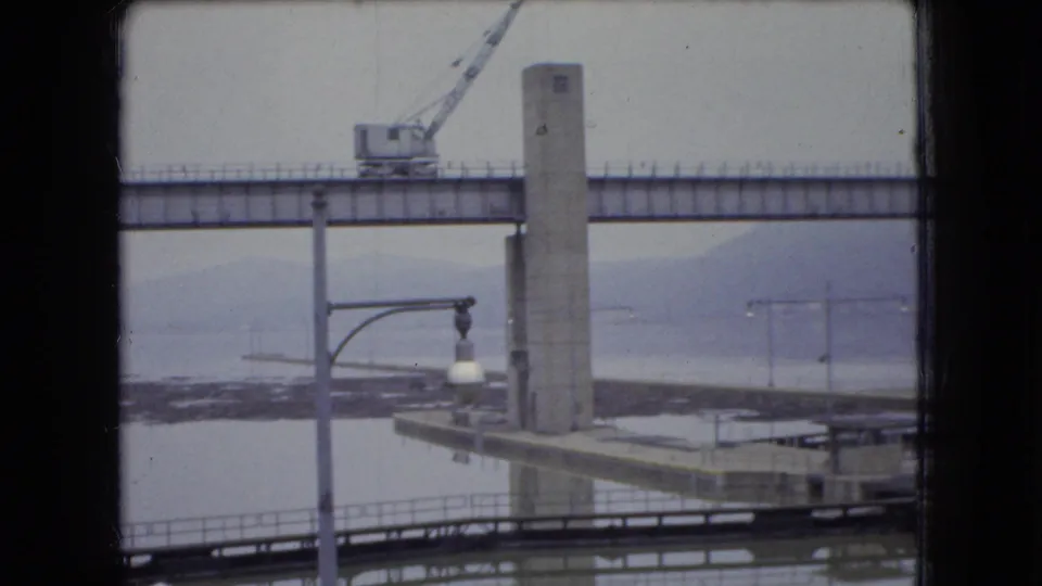 1966 Ohio 8mm frame showing an industrial crane on a high bridge-like span above a wide river, with a concrete support tower and shoreline railings visible through a window frame.