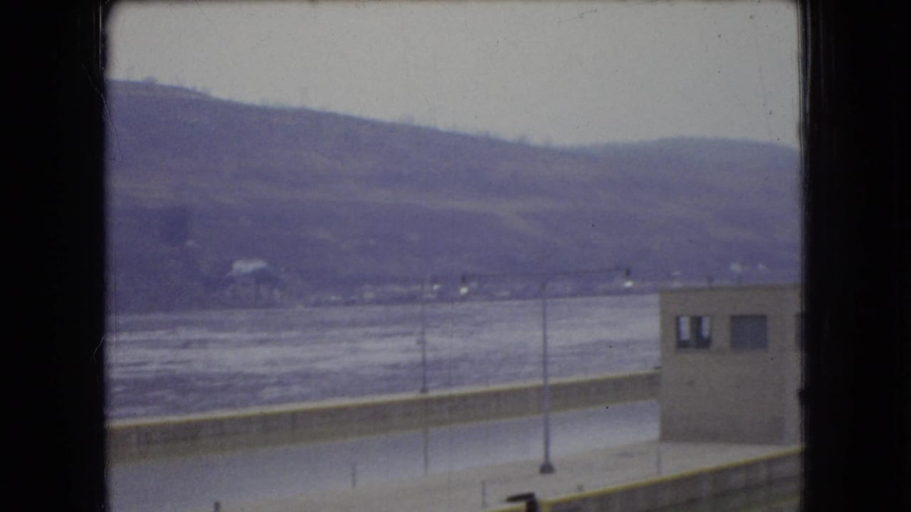 1966 Ohio home-movie frame looking across a broad river toward low hills, with a small utilitarian building and riverside pavement partly obscured by the window frame.