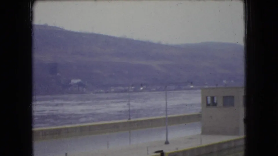 1966 Ohio home-movie frame looking across a broad river toward low hills, with a small utilitarian building and riverside pavement partly obscured by the window frame.