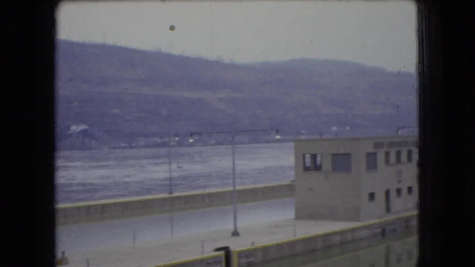 1966 Ohio 8mm view of a small control-like riverside building beside a wide river, with lamp posts, a low barrier, distant hills, and dark frame bars in the foreground.
