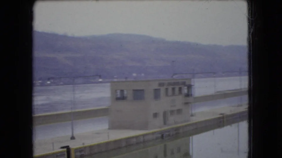 Final 1966 Ohio home-movie frame showing the riverside building, railing or walkway, water, distant hills, and heavy dark window-frame bars dividing the composition.