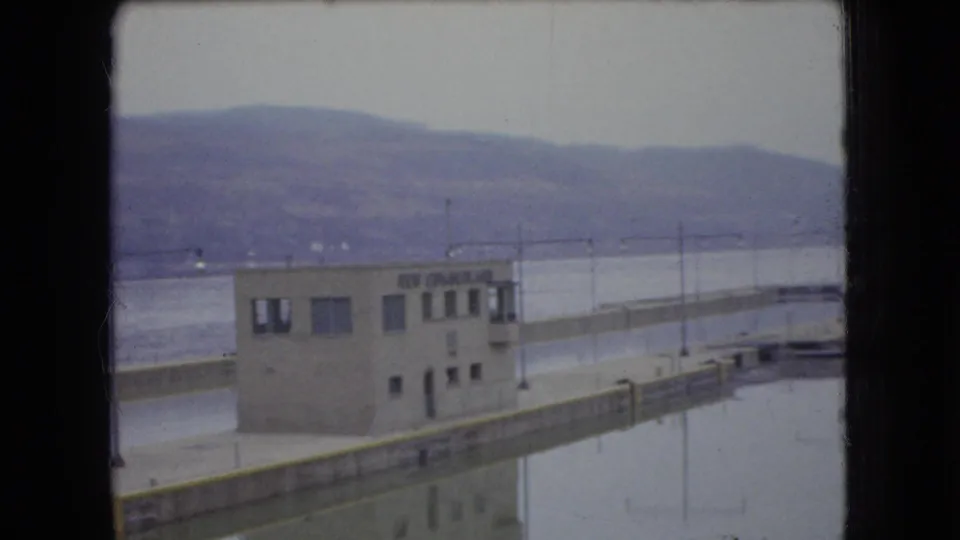 1966 Ohio 8mm frame showing a concrete dock or lock building beside still water, with a long pier wall and distant hills under an overcast sky.