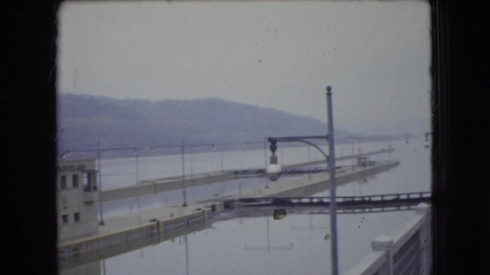 1966 Ohio 8mm frame looking across a quiet industrial dock or lock channel, with a lamp post in the foreground and hills fading into the cloudy background.