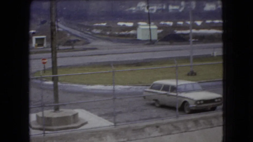 1966 Ohio home-movie frame showing a white station wagon near a chain-link fence, roadside signs, utility poles, and the waterway facility beyond.