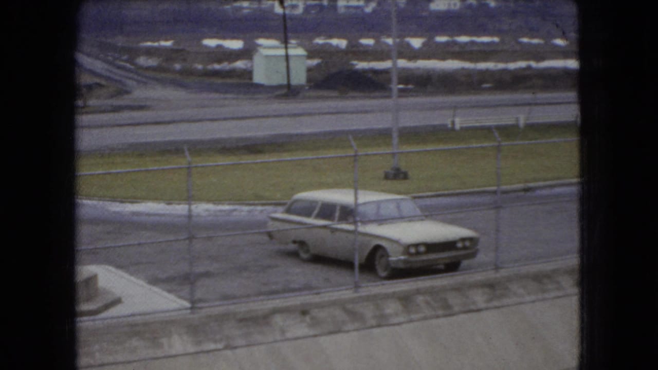 A 1966 Ohio home-movie frame shows a pale station wagon parked beyond a chain-link fence beside wet pavement and roadside grass.