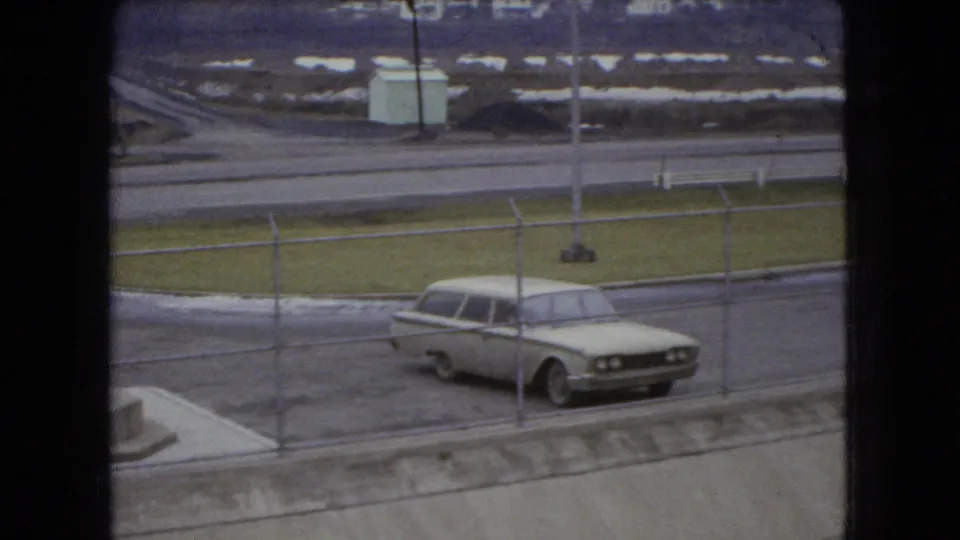 A 1966 Ohio home-movie frame shows a pale station wagon parked beyond a chain-link fence beside wet pavement and roadside grass.