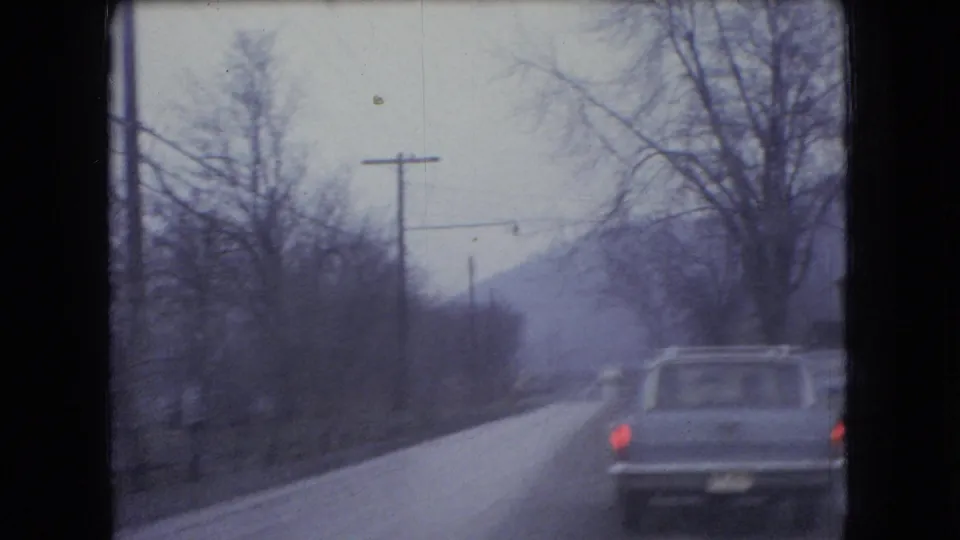 A car travels ahead on a rural Ohio road lined with bare trees and utility poles, recorded in soft, grainy 8mm footage from 1966.