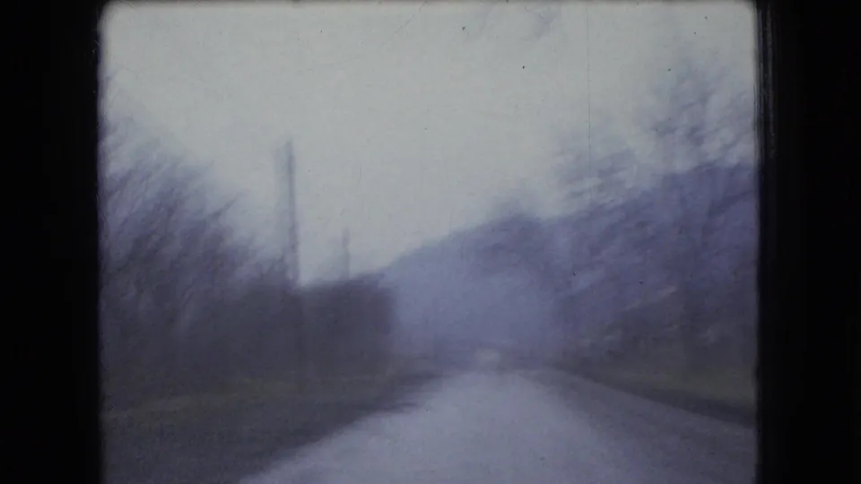 1966 Ohio 8mm frame from a moving car looking down a quiet rural road bordered by bare trees, utility poles, and low hills.