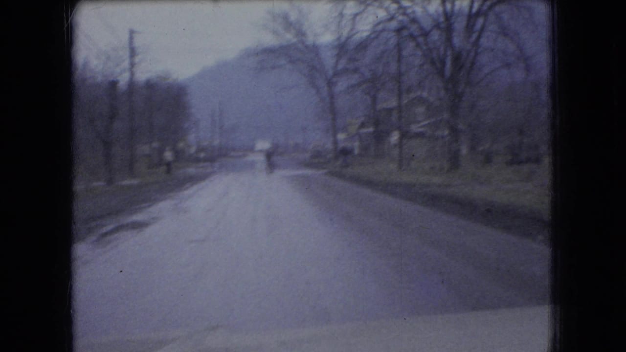 1966 Ohio home-movie frame showing a moving-car view along a small-town road with bare trees, utility poles, distant hills, and a vehicle ahead.