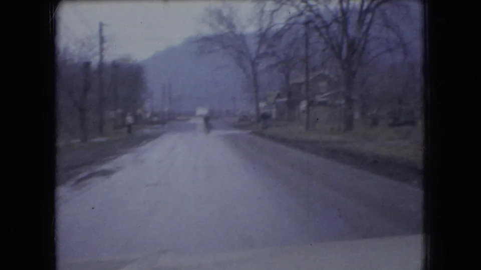1966 Ohio home-movie frame showing a moving-car view along a small-town road with bare trees, utility poles, distant hills, and a vehicle ahead.
