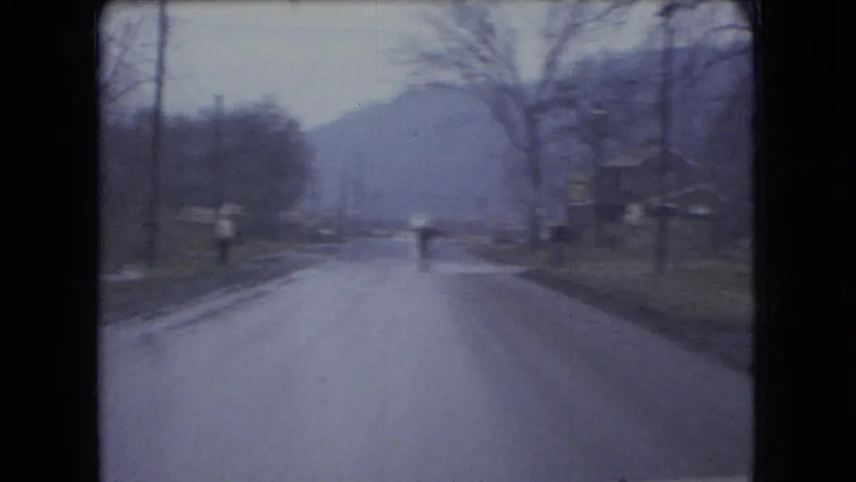 1966 Ohio 8mm frame looking down a quiet road where a distant person or roadside figure appears near the center, with bare trees and hills beyond.