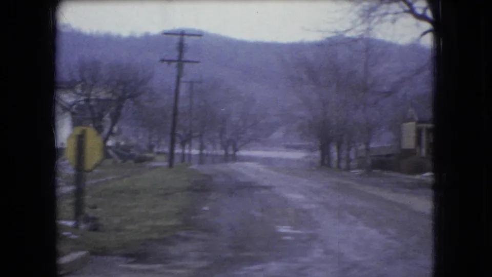 1966 Ohio home-movie frame from a moving car showing a roadside bend with a yellow sign, utility pole, bare trees, wet-looking pavement, and wooded hills.
