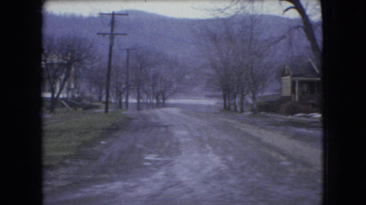 1966 Ohio 8mm frame looking down a slushy residential hill lined with bare trees, utility poles, and houses in winter conditions.
