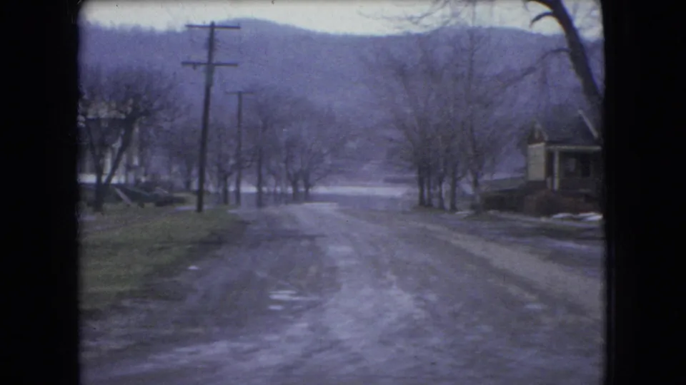 1966 Ohio 8mm frame looking down a slushy residential hill lined with bare trees, utility poles, and houses in winter conditions.