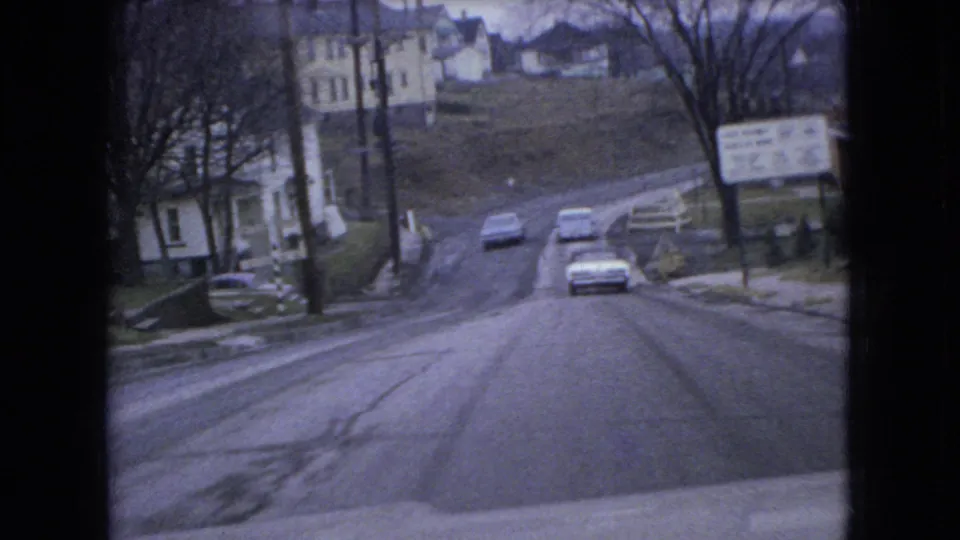 Cars move along a slushy downhill residential street in 1966 Ohio, passing houses, utility poles, bare trees, and a small roadside sign.