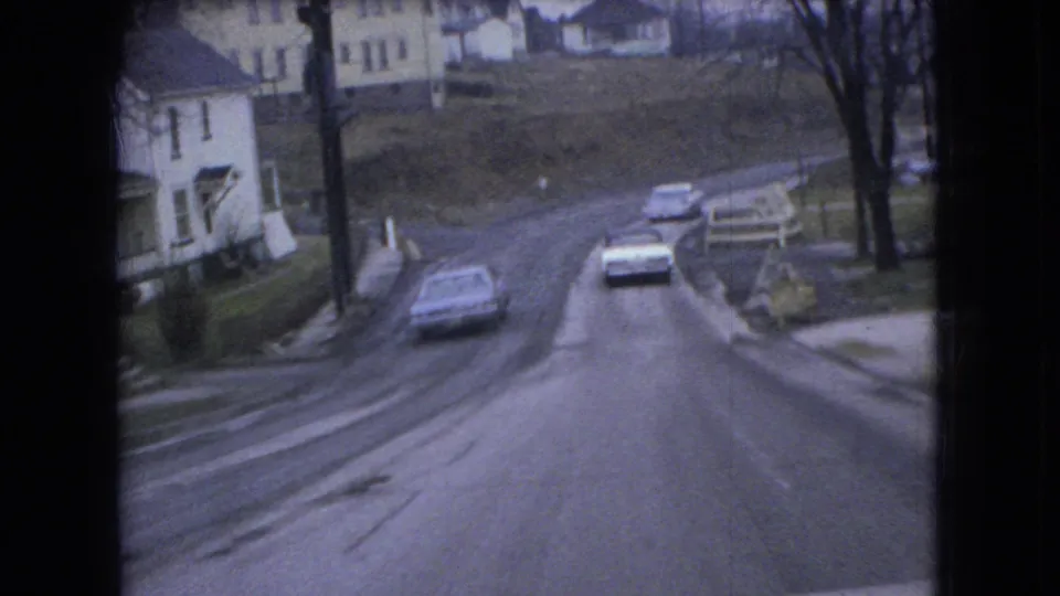 Two cars travel down an icy Ohio neighborhood road in a grainy 8mm frame, with houses, bare trees, and wet pavement visible around them.