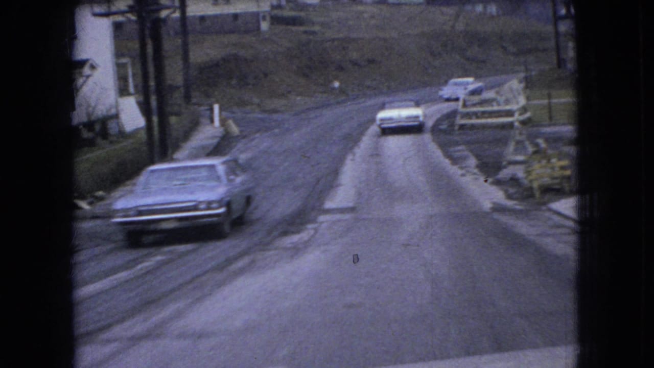 1966 Ohio 8mm frame showing older American sedans on a rough curving rural road with modest roadside houses and a hillside in view.