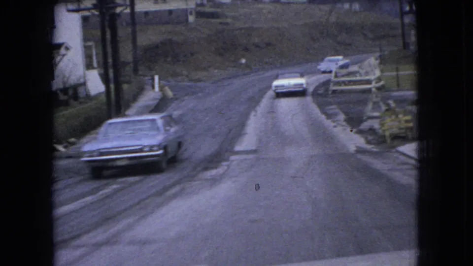 1966 Ohio 8mm frame showing older American sedans on a rough curving rural road with modest roadside houses and a hillside in view.