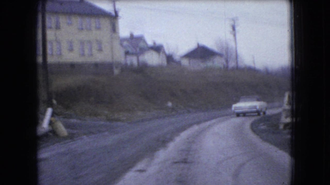 1966 Ohio 8mm frame with a white sedan rounding a curved rural road below a pale multi-window building and small houses.