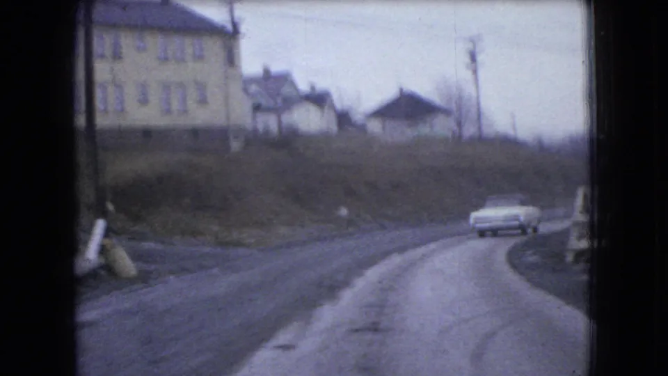 1966 Ohio 8mm frame with a white sedan rounding a curved rural road below a pale multi-window building and small houses.
