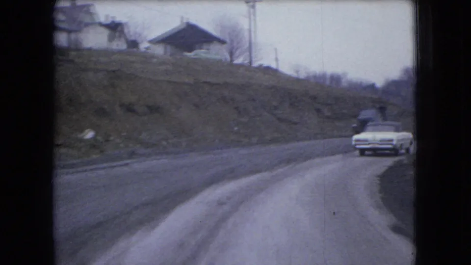 1966 Ohio 8mm frame showing a white sedan approaching along a curved road bordered by rough hillside ground and distant houses.