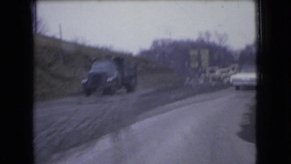 1966 Ohio 8mm frame showing a dark dump truck and a white sedan sharing a rough curving rural road near roadside buildings.