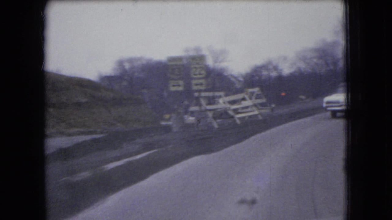 1966 Ohio 8mm frame from a moving vehicle showing a winding road, roadside signs, construction-style barriers, bare trees, and a white sedan ahead.