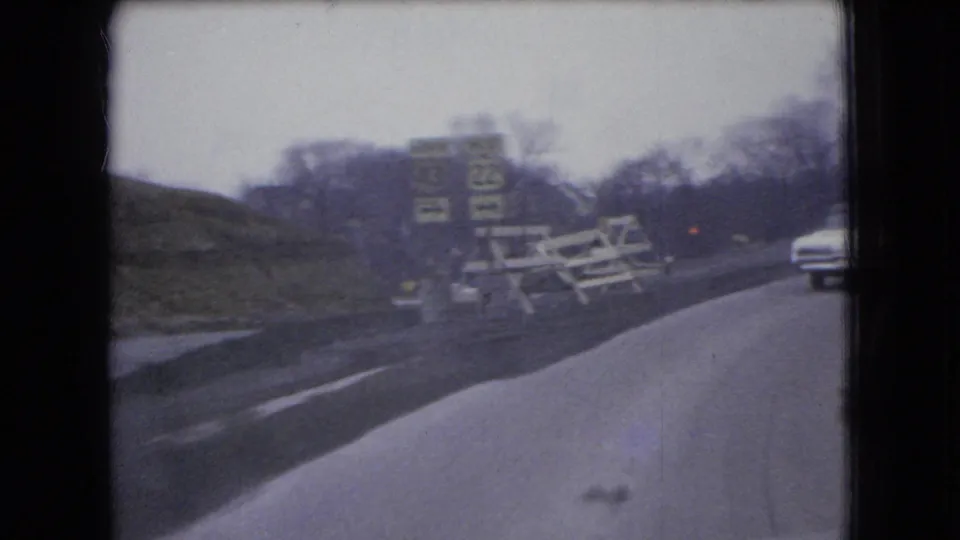 1966 Ohio 8mm frame from a moving vehicle showing a winding road, roadside signs, construction-style barriers, bare trees, and a white sedan ahead.
