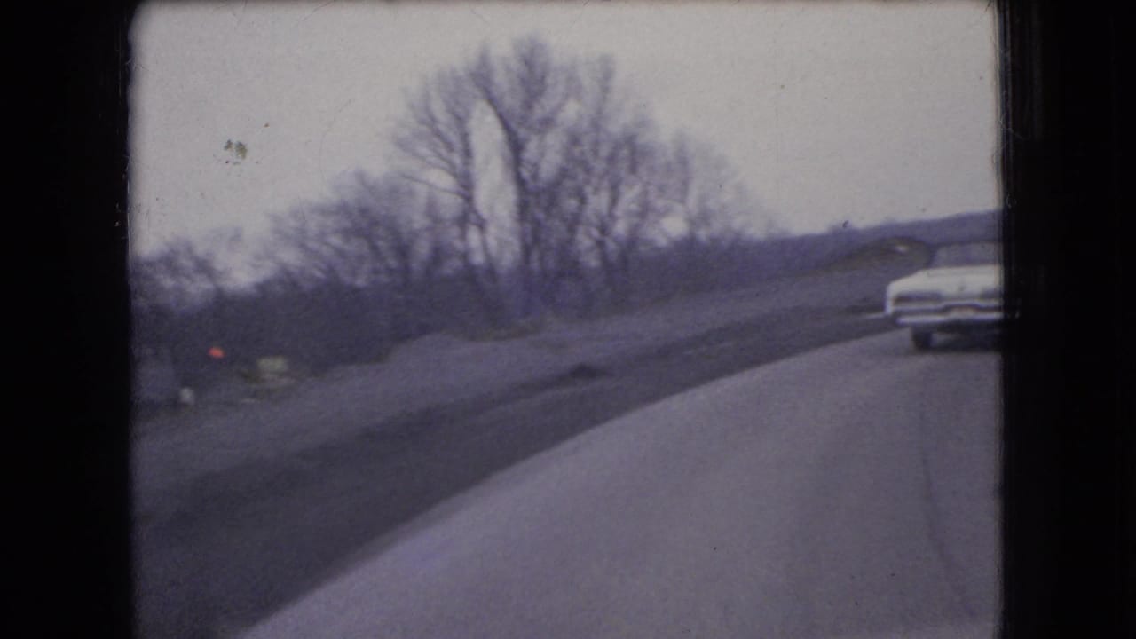 1966 Ohio 8mm frame showing a white sedan on a curving road bordered by bare trees and an open roadside embankment.