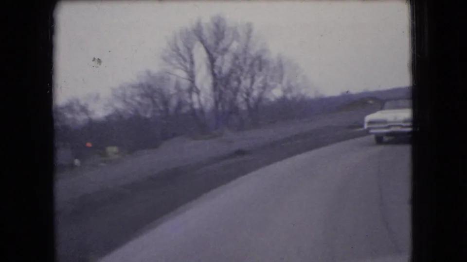 1966 Ohio 8mm frame showing a white sedan on a curving road bordered by bare trees and an open roadside embankment.