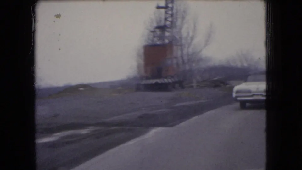 1966 Ohio 8mm frame from a moving car showing a roadside industrial structure or crane near a curving road and a white sedan ahead.