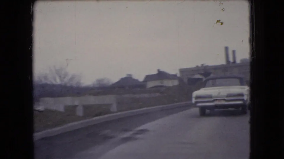 1966 Ohio 8mm frame showing a white sedan on the road with low industrial buildings, bare trees, barriers, and smokestacks in the background.