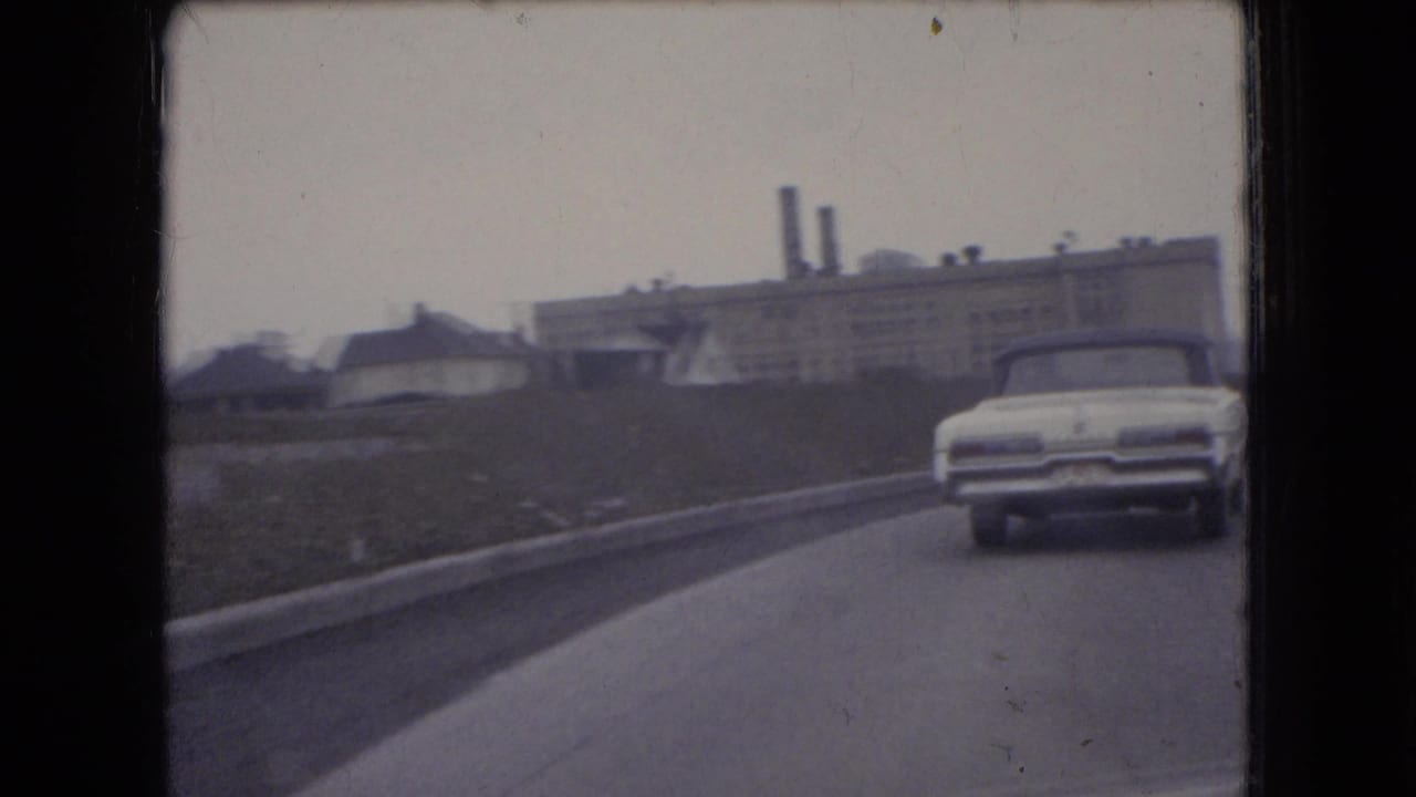 White mid-century car on a divided road near low industrial buildings and smokestacks.