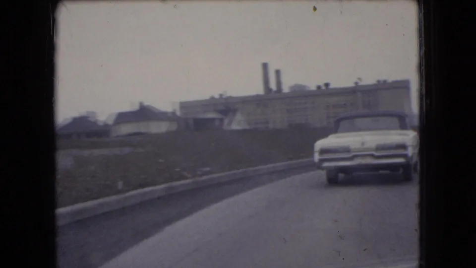 A white mid-century car drives ahead on an Ohio roadway in 1966, with low industrial buildings and twin smokestacks visible beyond the embankment.