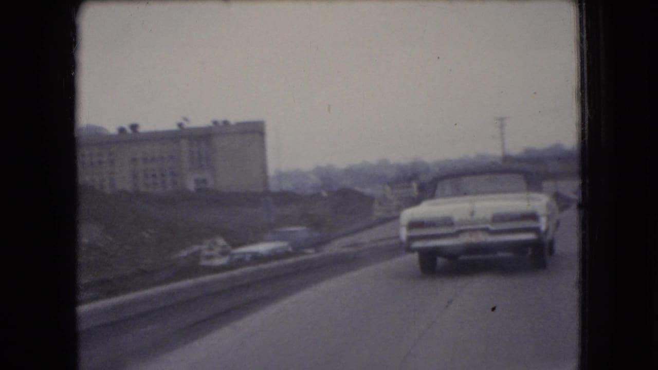 The moving-vehicle view follows a white car along an Ohio road in 1966, with an industrial building, roadside grading, and a utility pole in the background.