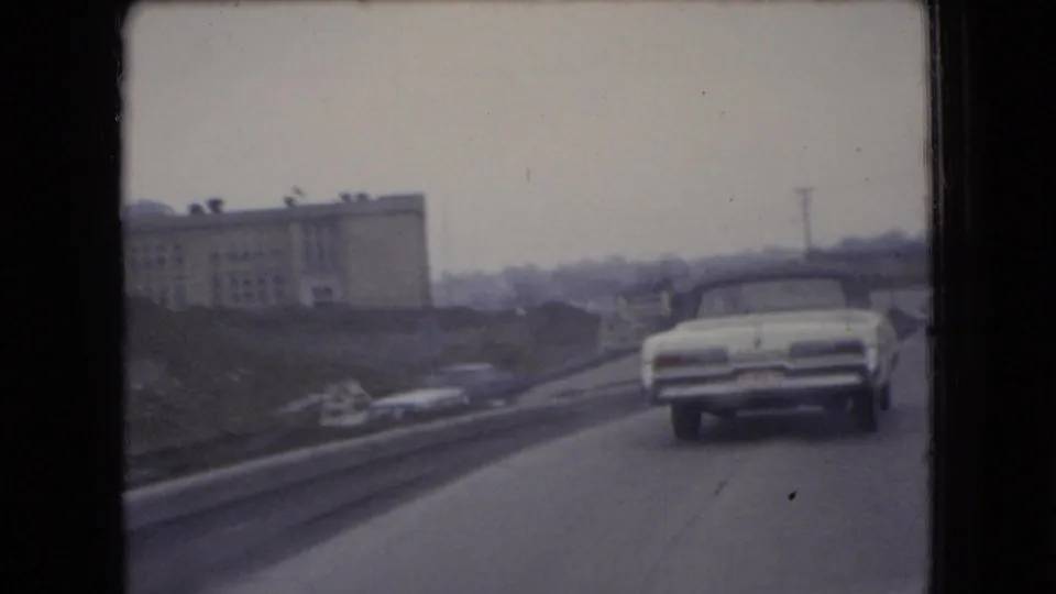 The moving-vehicle view follows a white car along an Ohio road in 1966, with an industrial building, roadside grading, and a utility pole in the background.