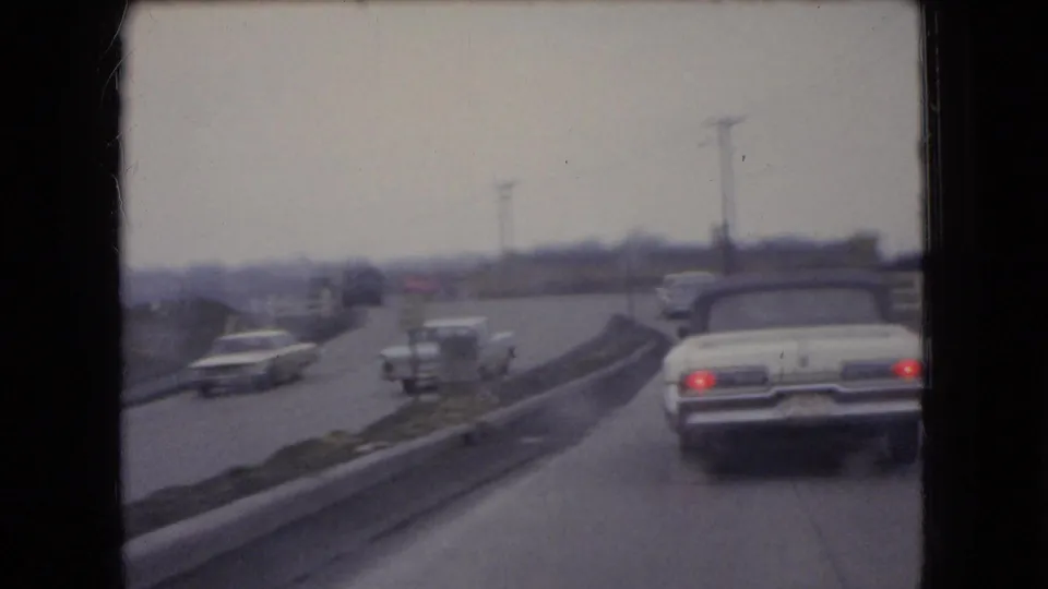 A rear view from a moving car shows a white convertible ahead on an Ohio divided road in 1966, with other mid-century cars traveling on the opposite side.