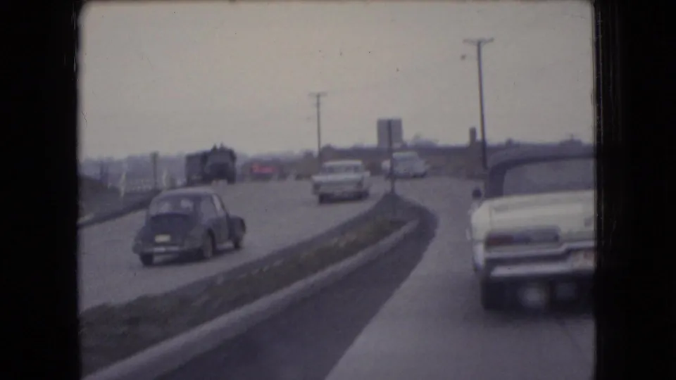 Several mid-century cars move along a divided Ohio roadway in 1966, including a dark sedan and a white car, with utility poles and distant industrial structures in view.