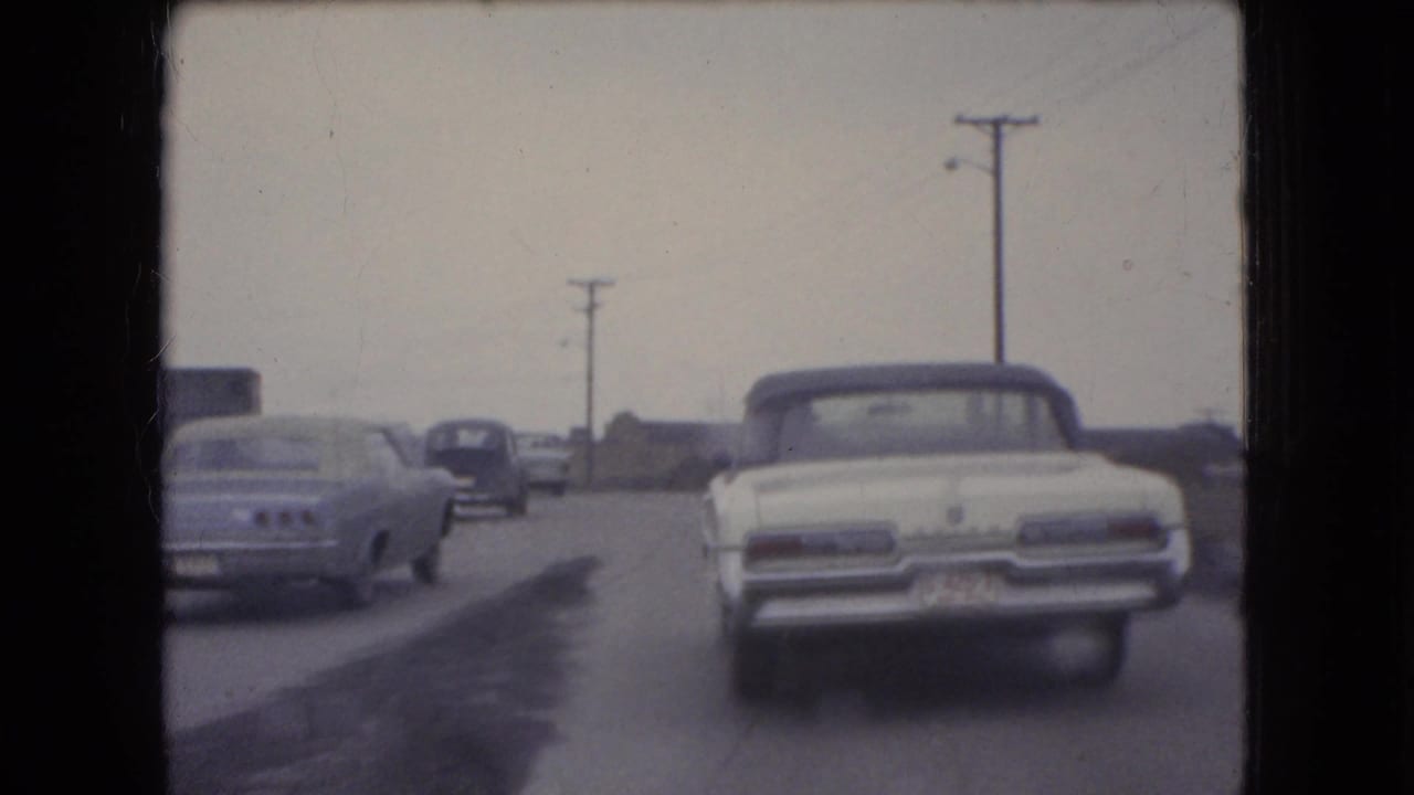 White convertible ahead of the camera with several cars on a road lined by utility poles.