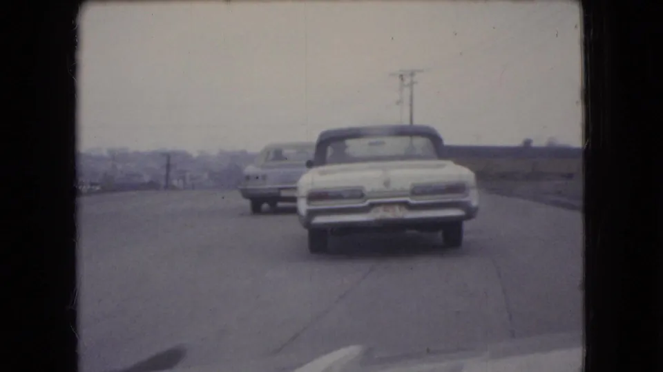 1966 Ohio 8mm frame showing the rear of a white convertible and another period car on a paved road with open roadside land and utility poles.