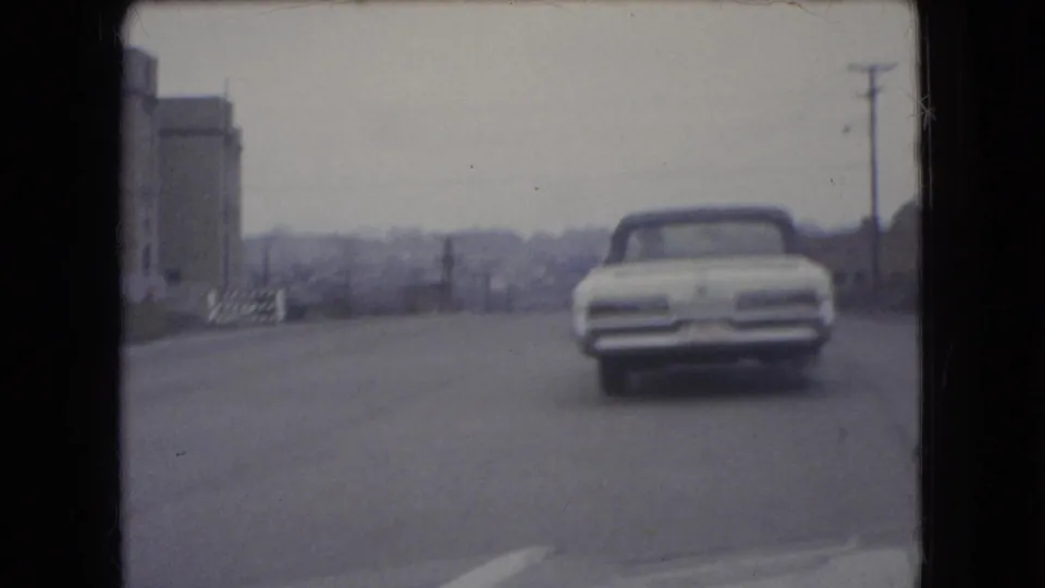 1966 Ohio 8mm frame showing a white convertible driving away on a two-lane road near a large roadside structure, utility poles, and distant trees.