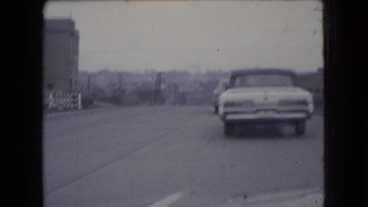 In 1966 Ohio 8mm footage, a white mid-century car drives away from the camera on a paved road with buildings and utility poles under an overcast sky.