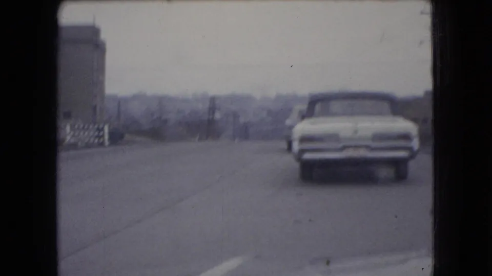 In 1966 Ohio 8mm footage, a white mid-century car drives away from the camera on a paved road with buildings and utility poles under an overcast sky.