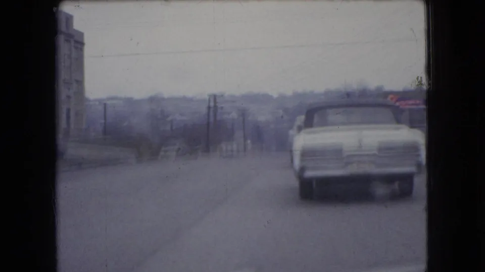 The white 1960s car continues away along an Ohio roadway, with a building at left, utility poles, and a muted winter landscape in the background.