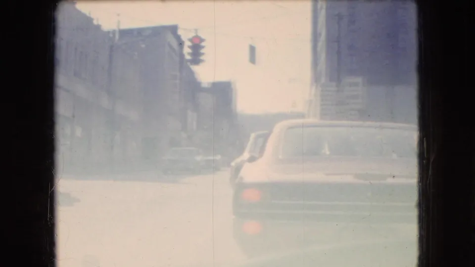 Soft 1966 Ohio street view through glass, with older cars ahead, building facades along the road, and a hanging traffic signal in the distance.