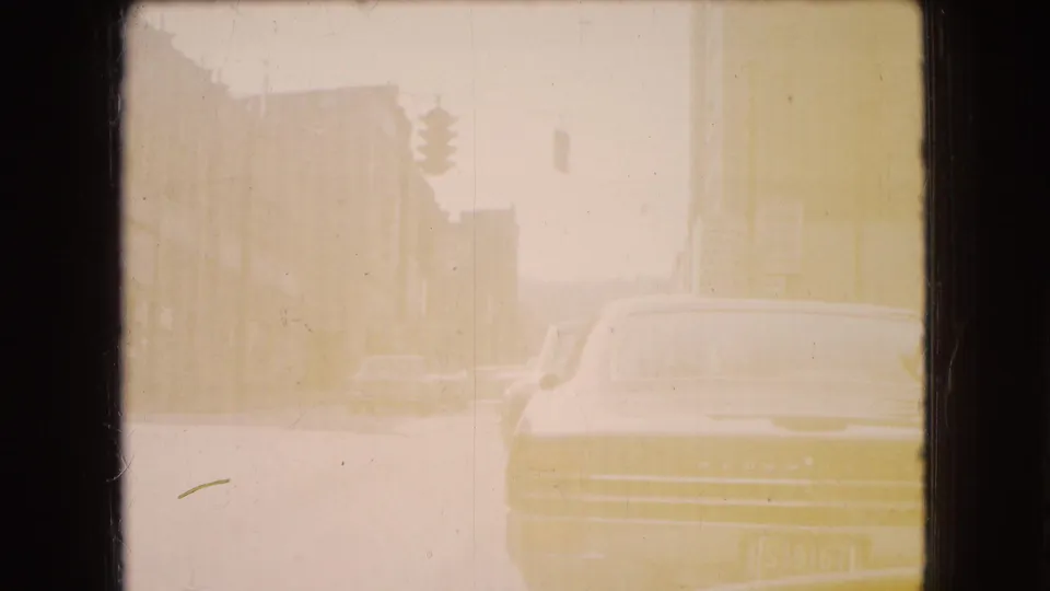 Overexposed yellow-toned 8mm frame of an Ohio city street, showing older cars, building edges, and a suspended traffic signal through a hazy window view.
